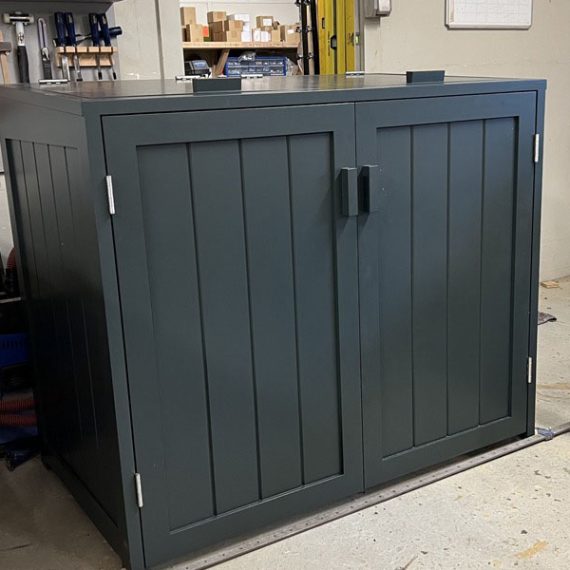 Three-quarter angle view of a bespoke double wheelie bin store crafted from Accoya wood and painted deep charcoal grey, made for London townhouse sitting on a workshop floor.