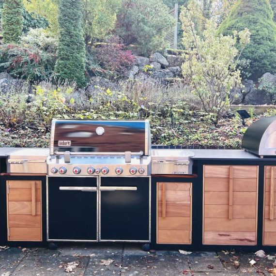 Bespoke outdoor kitchen featuring handcrafted hardwood cabinetry with integrated wood-handled drawers, a premium built-in gas BBQ, and a stainless steel Igneus pizza oven on a dark stone worktop.
