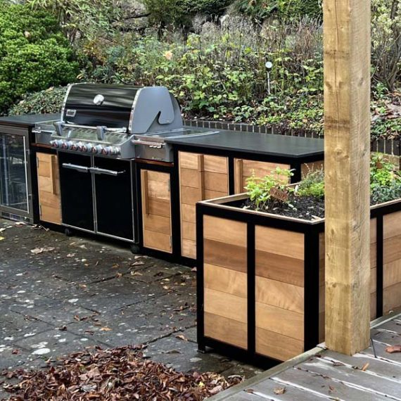 Oak long lasting L-shaped outdoor kitchen with custom herb planter and metal bracing detail and fridge.