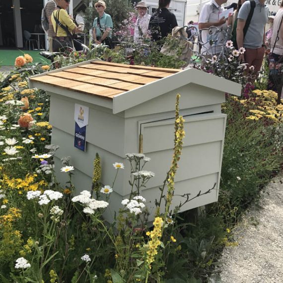 Bespoke wooden outdoor storage cabinet painted sage green with a cedar shingle roof, displayed in a wildflower garden bed at the RHS Chelsea Flower Show.