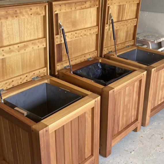 Bespoke hardwood outdoor litter bins with heavy-duty gas struts holding the top lids open, showing internal metal bin liners inside a joinery workshop.