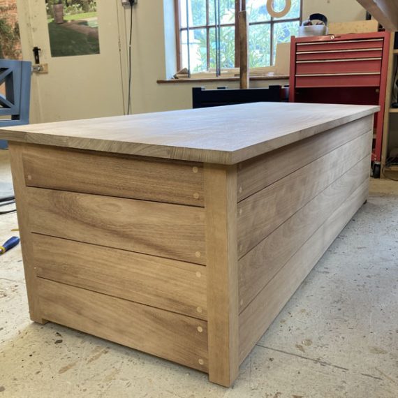 Long outdoor oak pool storage box handcrafted from solid hardwood timber slats with visible dowel joinery details, sitting on a workshop floor.