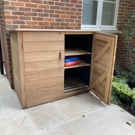 Bespoke hardwood outdoor storage cupboard with an open slatted ledger door showing internal shelves, placed against a red brick wall on a stone patio.