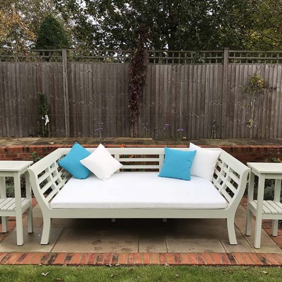 Bespoke wooden garden day bed painted off-white with a slatted backrest and slatted flared armrests, featuring a large white base cushion, turquoise pillows, and two matching tall, two-tier slatted wooden side tables.