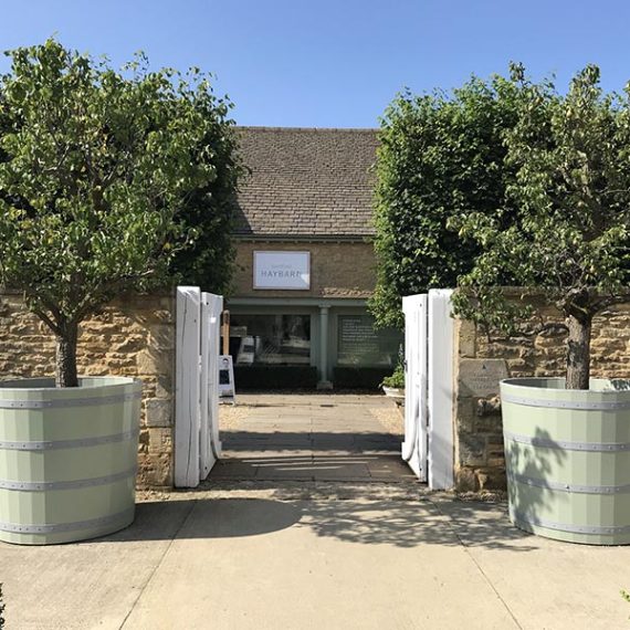 Pair of extra-large sage green Accoya barrel planters with decorative metal bands and mature trees framing an entrance at Daylesford Organic in the Cotswolds.