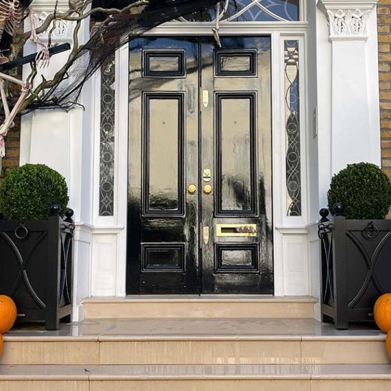 Pair of bespoke black Regency-style Accoya wooden planters with decorative panels and round topiary, framing the entrance of a private London townhouse.