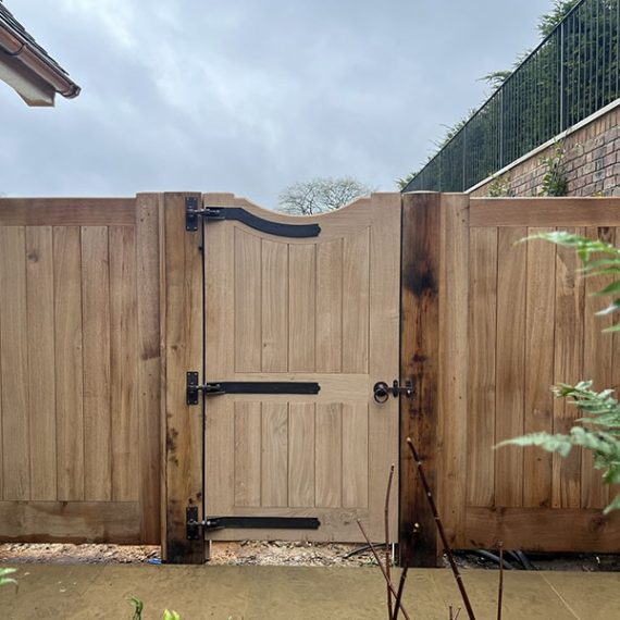 A luxury bespoke hardwood pedestrian gate with a dipped swan-neck top, flanked by solid timber side fencing panels and oak posts.