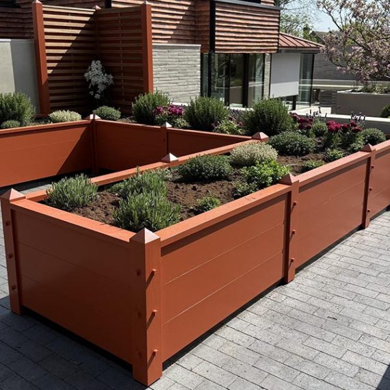 Bespoke made-to-order L-shaped raised timber beds handcrafted by Oxford Planters with a terracotta-orange finish, horizontal paneling, and pyramid finial posts on a contemporary stone paved terrace.