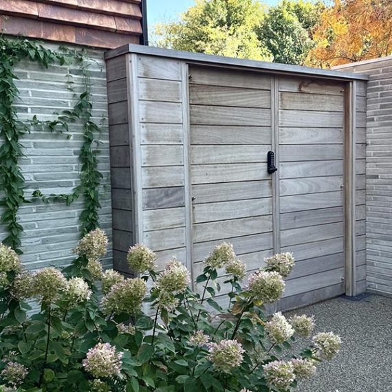 A flush-mounted wooden garden gate with a silver-grey patina and a mechanical lock, recessed into a brick wall