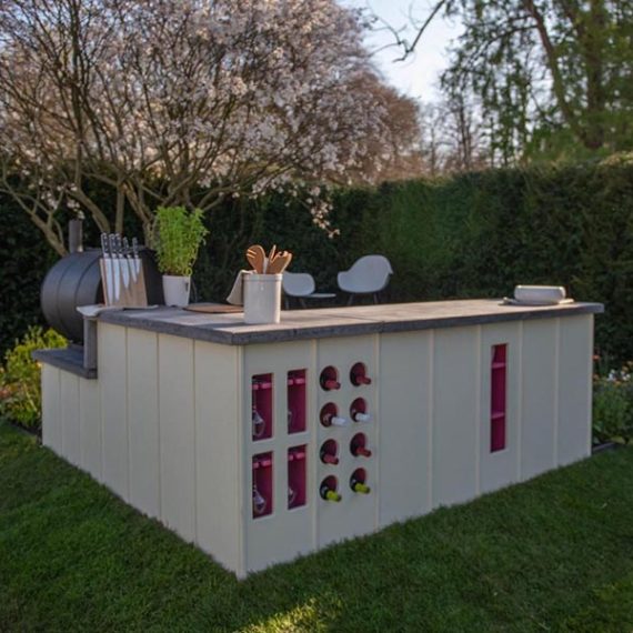 Bespoke outdoor kitchen featuring cream fluted panels, an integrated wine storage rack, and a concrete worktop set against a landscaped garden hedge.