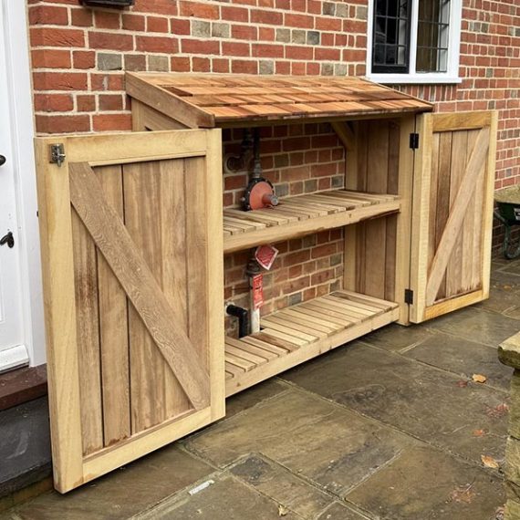 Bespoke hardwood outdoor storage cabinet with a cedar shingle roof, handmade in the UK with slatted shelving to neatly cover exterior utility pipes fixed to a brick wall.