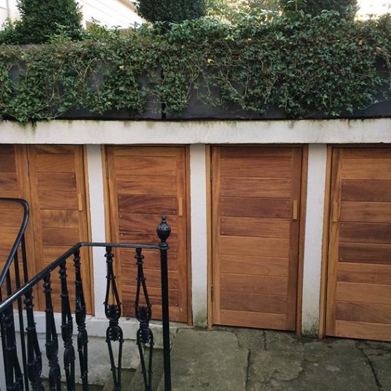 Four bespoke integral oak outdoor storage doors crafted from solid natural oak, installed flush within a concrete retaining wall underneath a garden terrace.