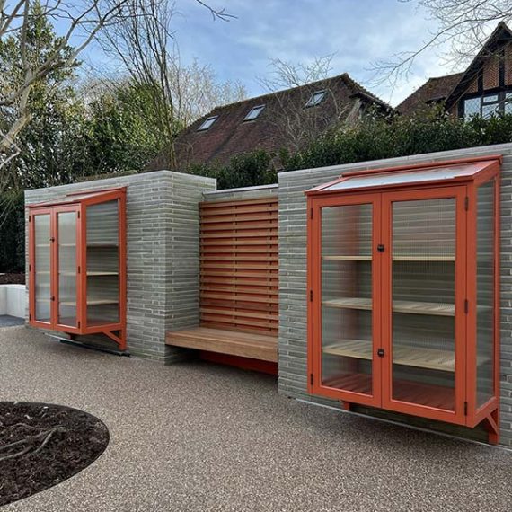 Two bespoke wall-mounted accoya cold frames painted terracotta orange with fluted glass doors, flanking a matching wooden slatted garden bench built into a brick wall.