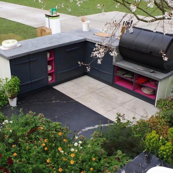 Bespoke L-shaped outdoor kitchen crafted with dark blue cabinetry, vibrant pink accent shelving, and a large black barrel BBQ smoker integrated into concrete countertops.