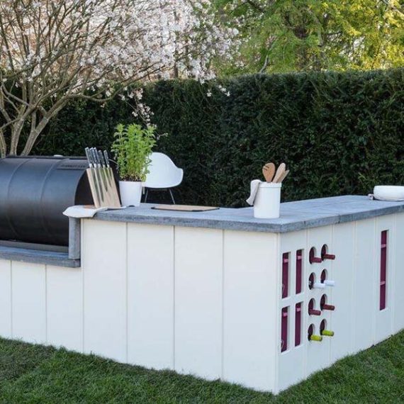 Rear view of a bespoke L-shaped outdoor kitchen featuring white fluted vertical cladding, a built-in wine rack with pink accents, and an integrated black barrel smoker on a lawn.