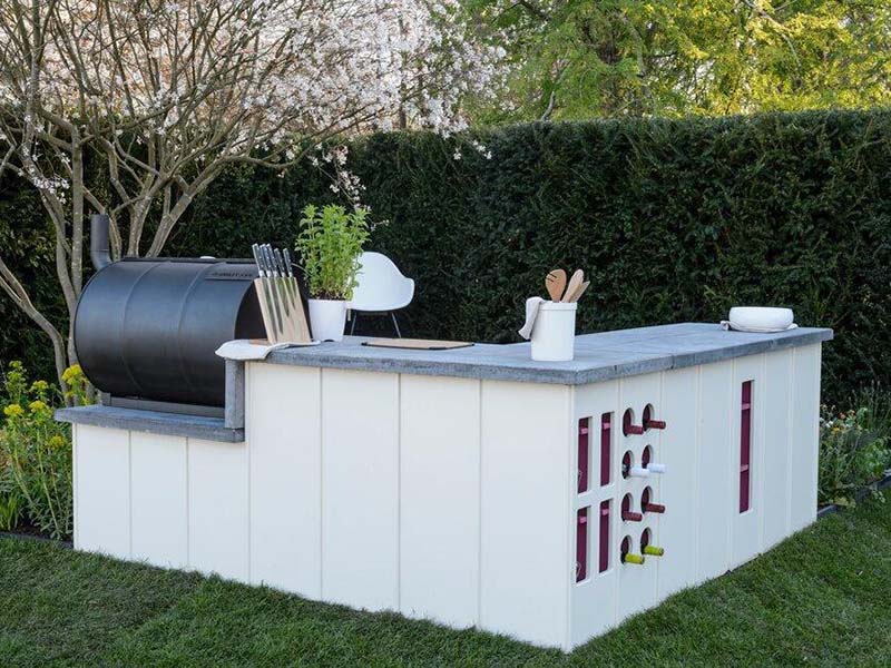 Fluted Outdoor Bespoke Kitchen with Wine Holder Rear view of a bespoke L-shaped outdoor kitchen featuring white fluted vertical cladding, a built-in wine rack with pink accents, and an integrated black barrel smoker on a lawn.