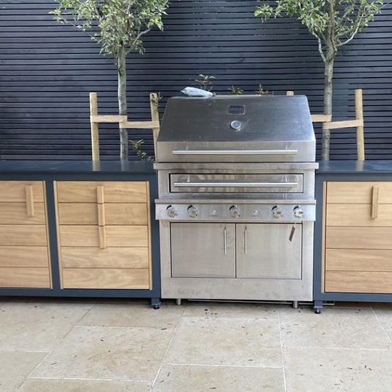 Luxury bespoke outdoor oak kitchen configuration featuring a large built-in stainless steel gas BBQ grill integrated between dark grey cabinets with natural oak panels.