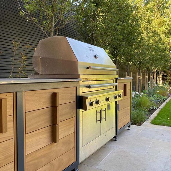 Side view of a luxury bespoke outdoor kitchen with oak and dark grey metal framing installed on a stone paved patio.