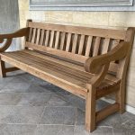 Extra-long heavy-section natural oak garden bench with classical scrolled armrests set against a stone masonry wall