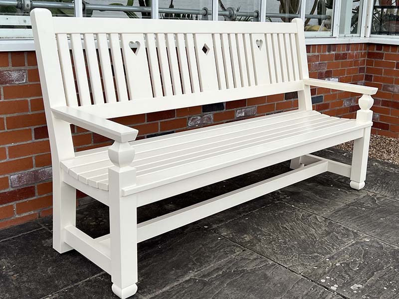 A bespoke National Trust commissioned Mawson garden bench painted finish, featuring distinctive hand-carved heart and diamond backrest details against a red brick wall.