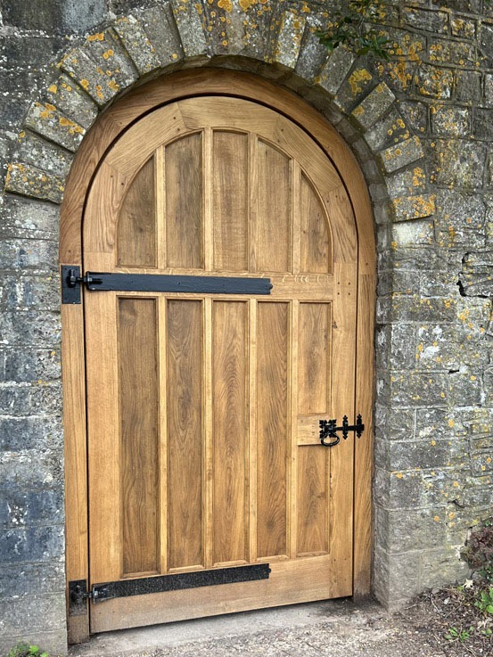 Bespoke Mawson-Inspired Heritage Oak Gate Bespoke heritage English hardwood oak gate replication featuring a soft segmental arch top, vertical tongue and groove timber boarding, and hand-forged black iron strap hinges set into a historic stone wall archway.