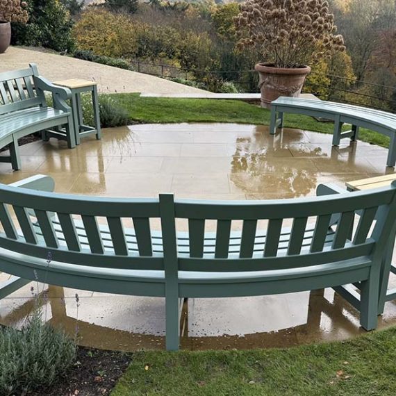 Bespoke curved wooden garden benches painted in sage green, arranged in a circular seating area on a flagstone patio overlooking a valley.