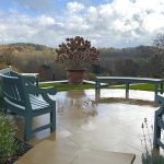 Bespoke curved hardwood garden benches painted sage green on a circular stone patio, looking out over a wide valley view.