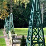 Row of sage green painted timber garden tuteurs with ball finials along a lawn pathway in formal estate grounds.